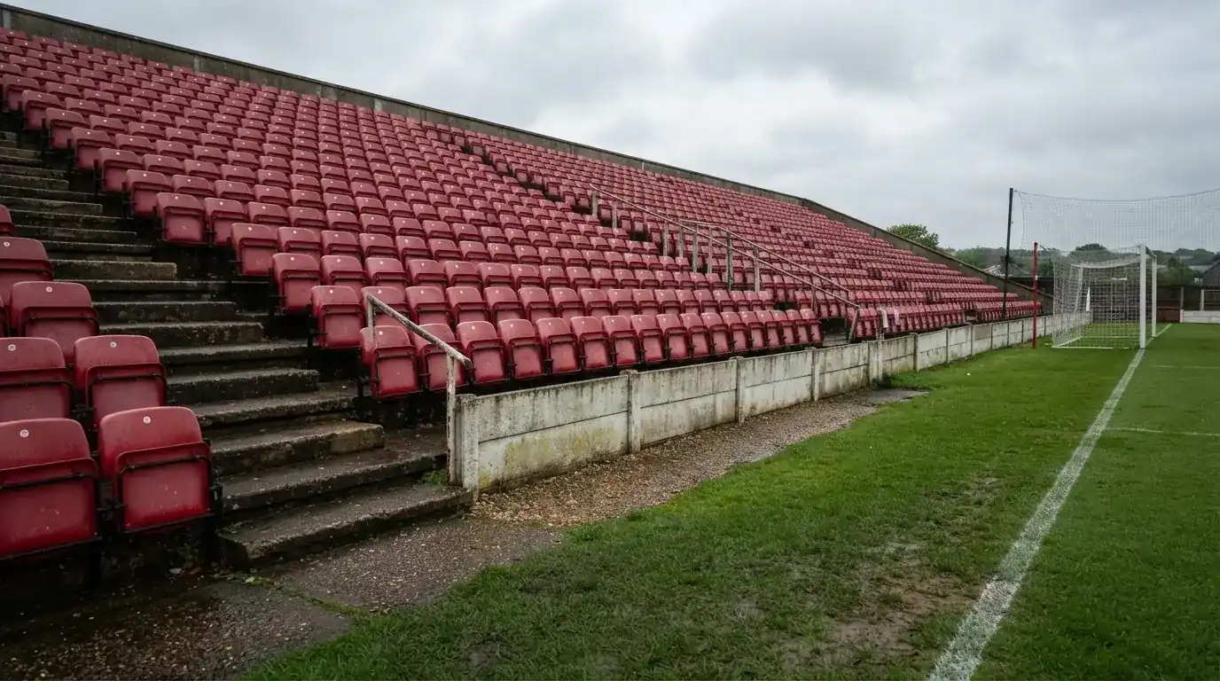 Rzędy pustych plastikowych krzesełek małego stadionu widziane z poziomu murawy pod pochmurnym niebem.