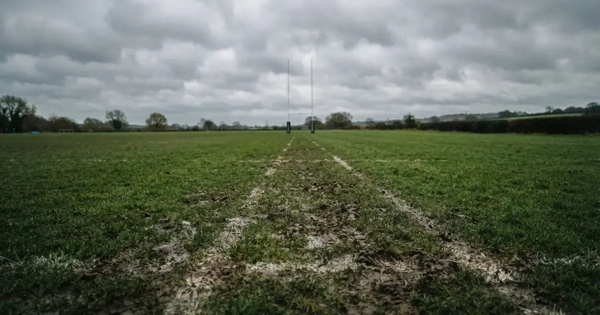 Boisko do rugby ze słupami bramki w oddali pod pochmurnym niebem, mokra trawa i ślady butów.