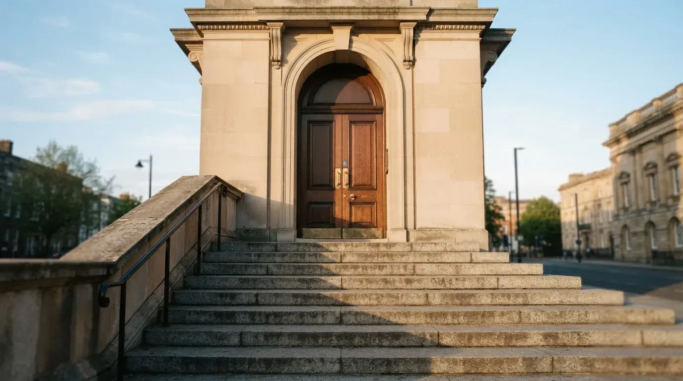 Granitowe schody prowadzące do wysokich drewnianych drzwi budynku w porannym świetle, pusta balustrada.